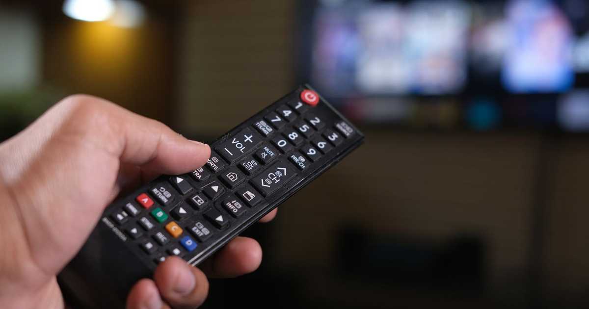 Pessoa segurando controle remoto com tela de TV desfocada ao fundo