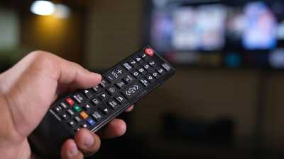Pessoa segurando controle remoto com tela de TV desfocada ao fundo