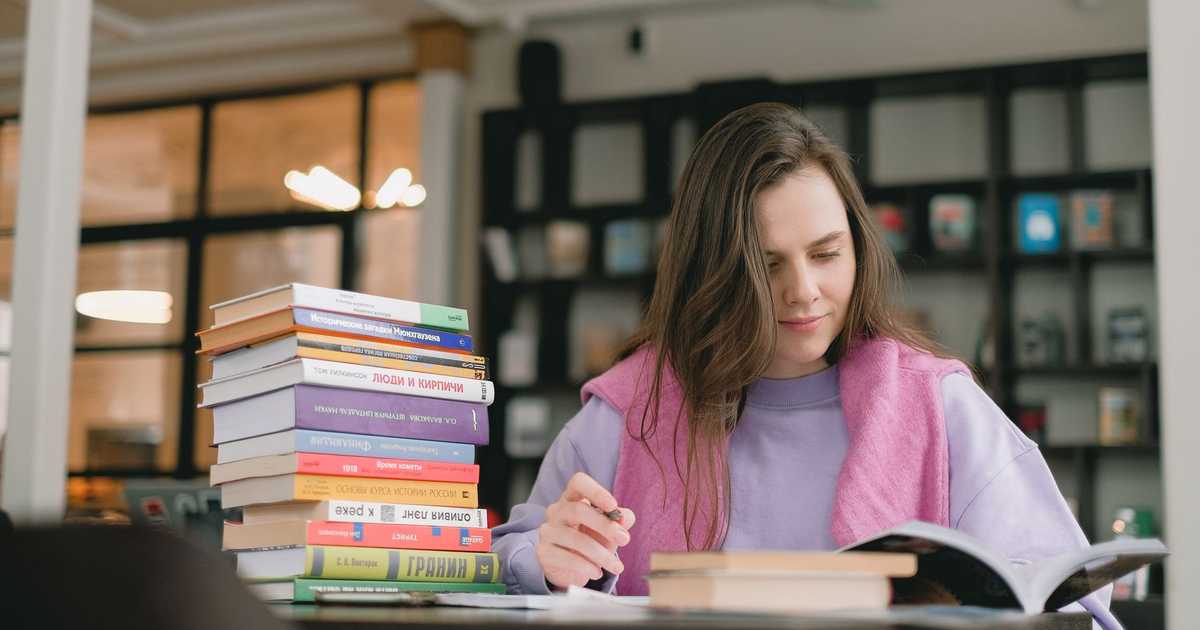 Jovem estudando intensamente em biblioteca, cercada de livros e anotações