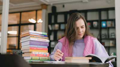 Jovem estudando intensamente em biblioteca, cercada de livros e anotações