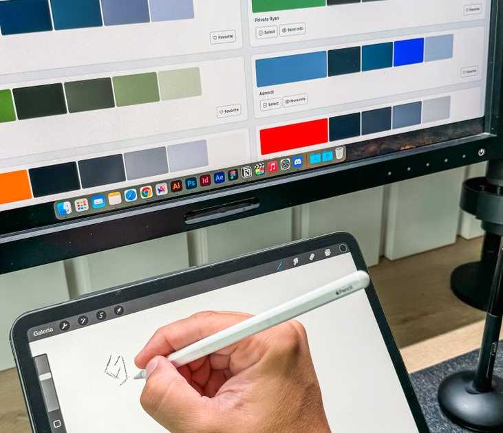Designer usando caneta digital em tablet com paleta de cores exibida em monitor